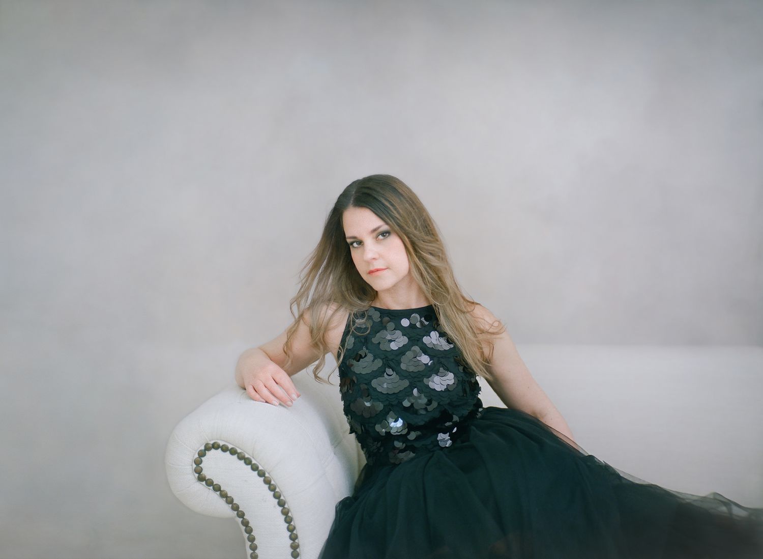 Model in black tulle gown poses on vintage chaise lounge against grey backdrop.