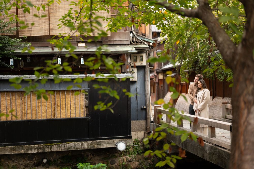 Romantic couple standing on a wooden bridge surrounded by lush green trees in Kyoto, Japan.