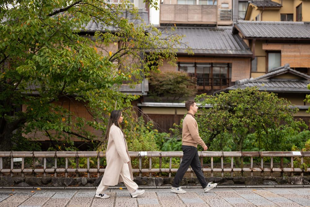 Couple walking side by side along a stone bridge with traditional Japanese architecture in background.