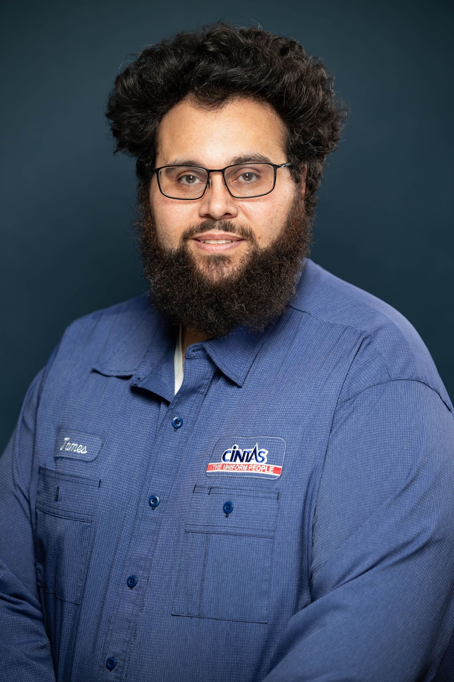 A person in a blue work uniform smiles at the camera while wearing glasses and sporting a full dark beard.