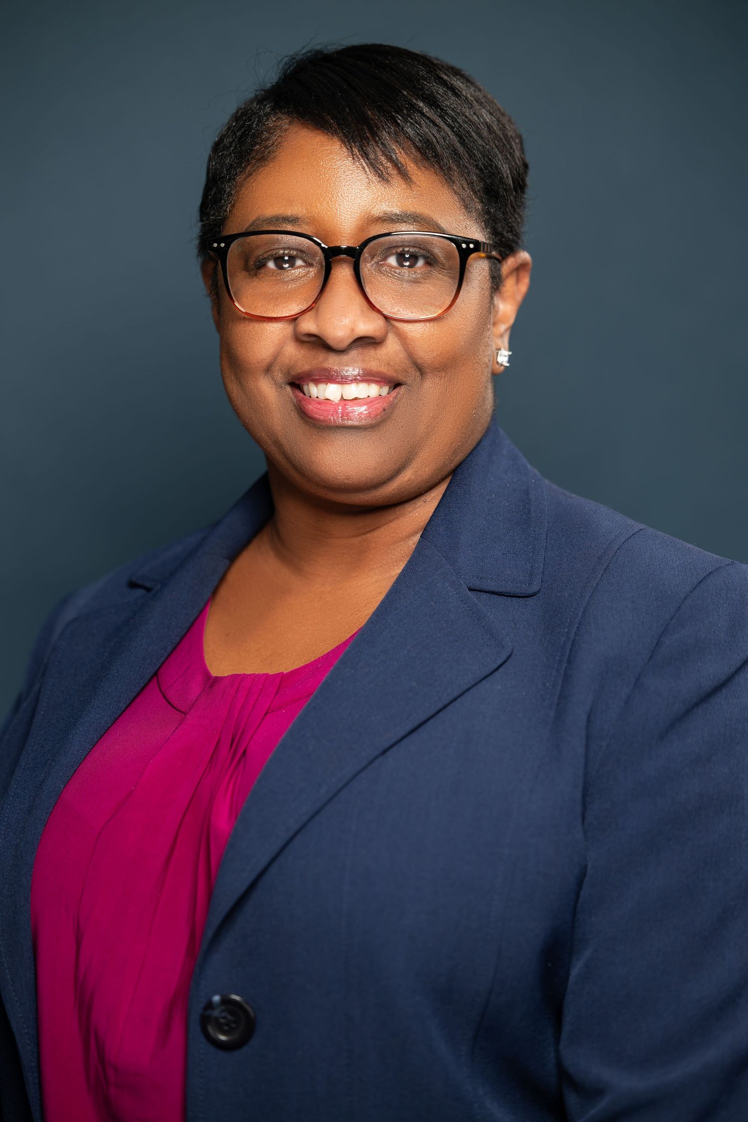 Professional headshot portrait showing person in navy blazer and magenta top against dark background with warm smile.