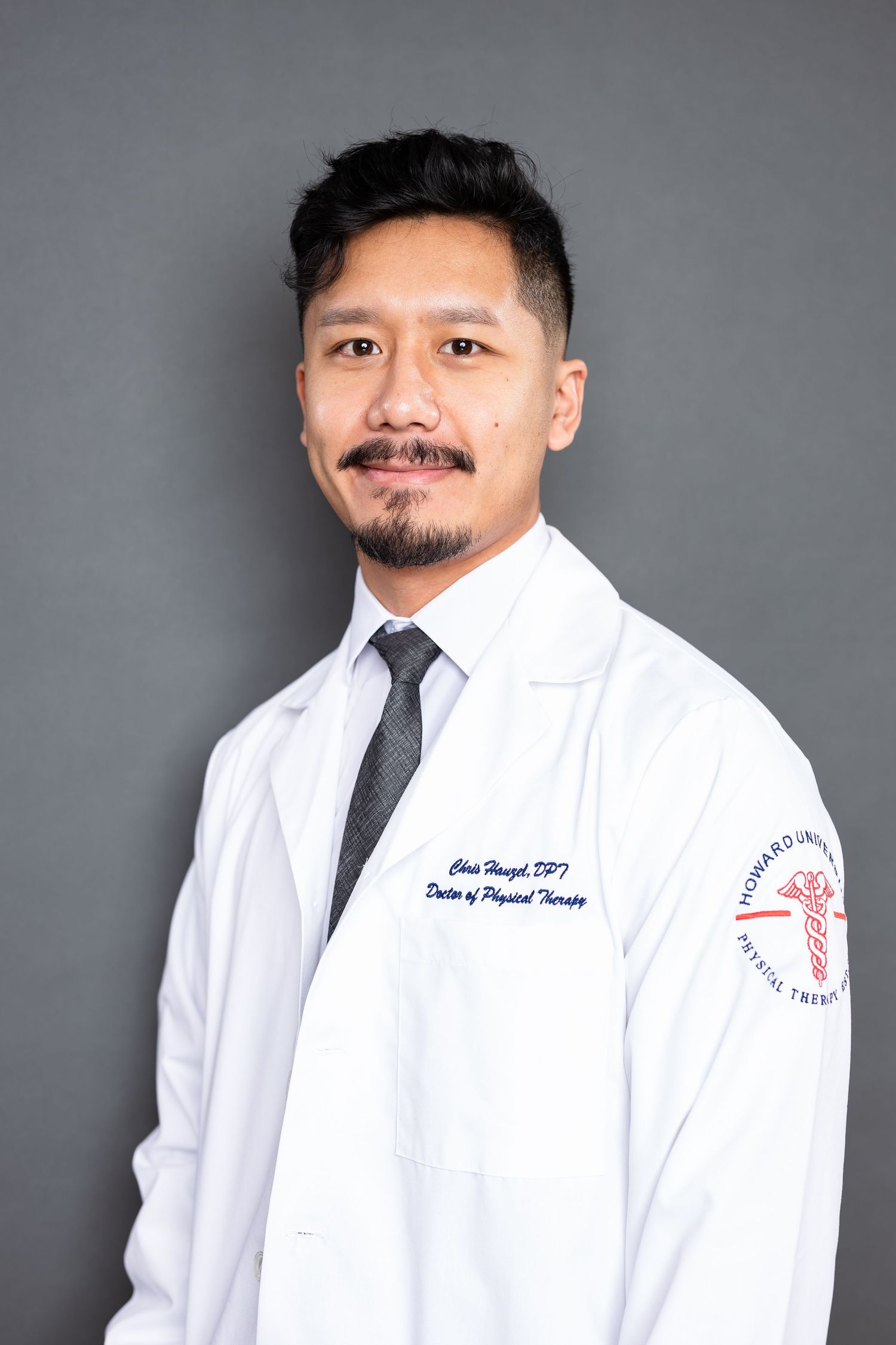 Healthcare professional in white coat and gray tie poses for portrait against gray background.