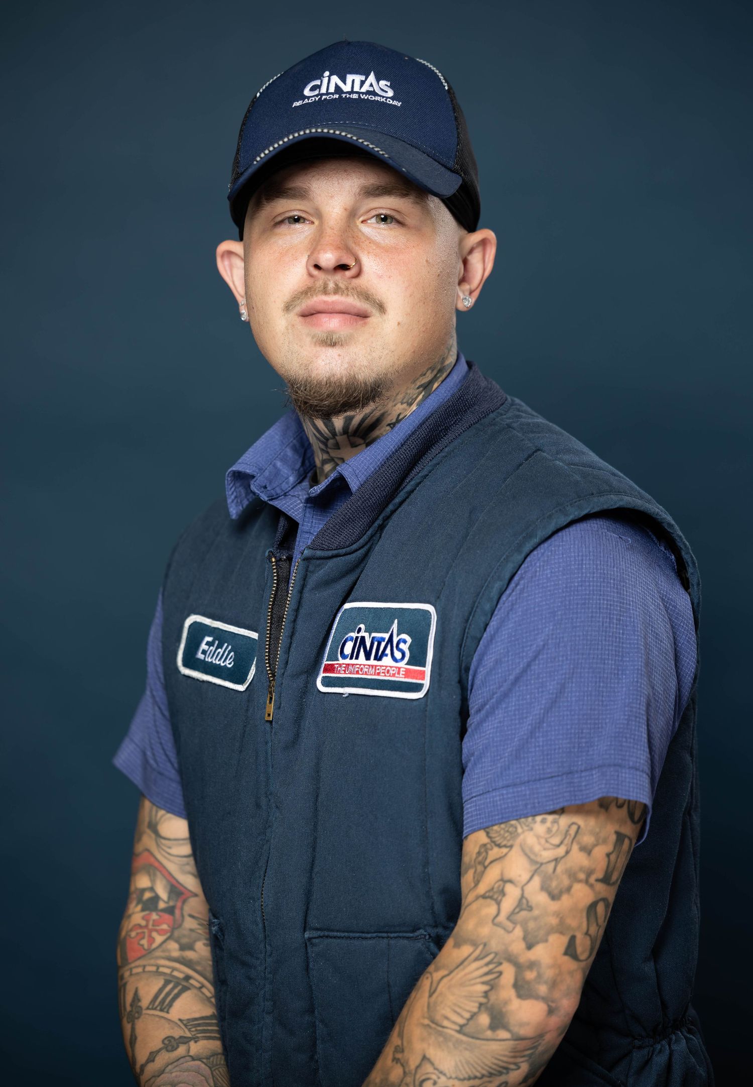 Person in navy blue baseball cap and vest showing tattoos on arms against dark background.