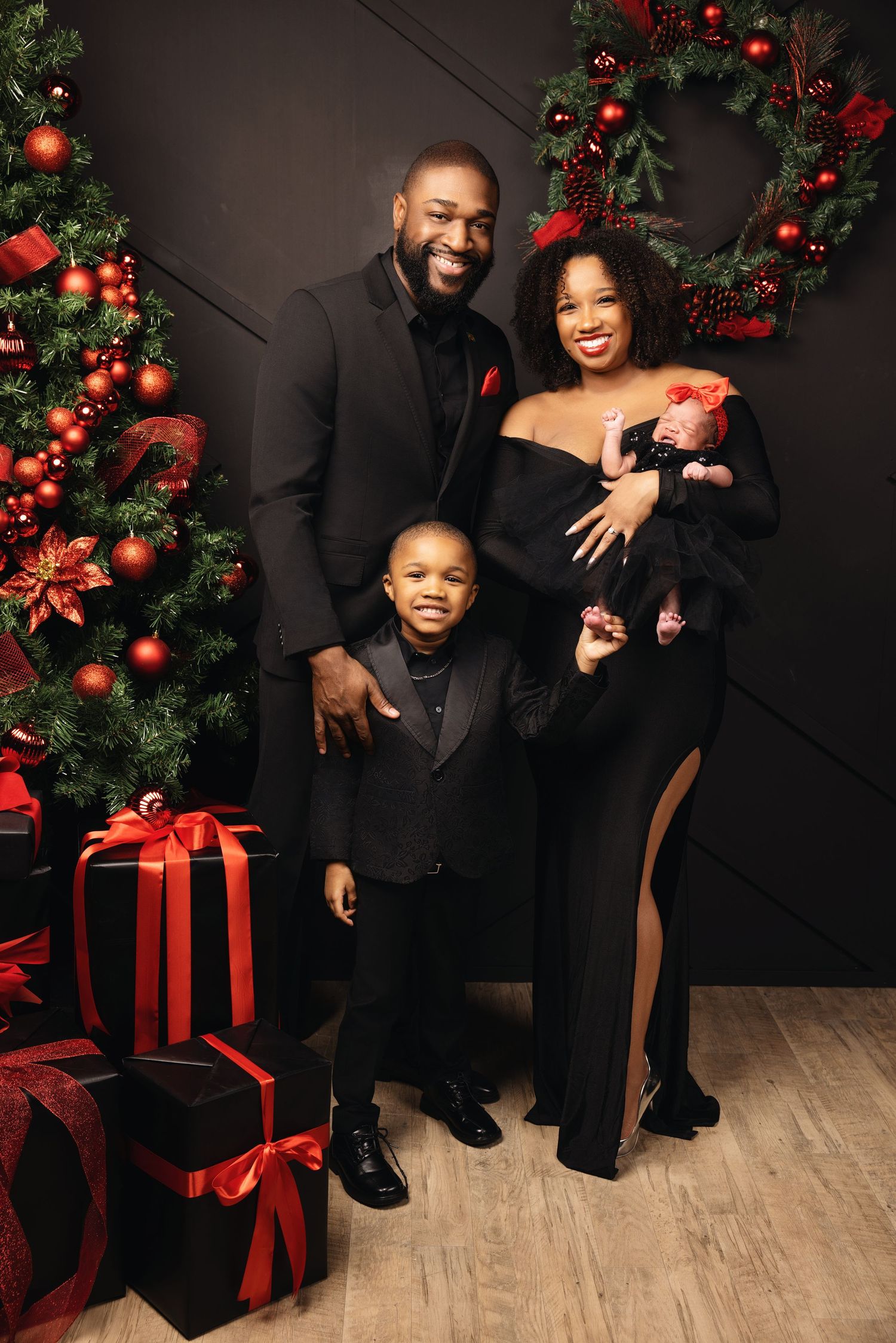 A family poses together in formal black attire against a festive Christmas backdrop with wreaths and wrapped presents.