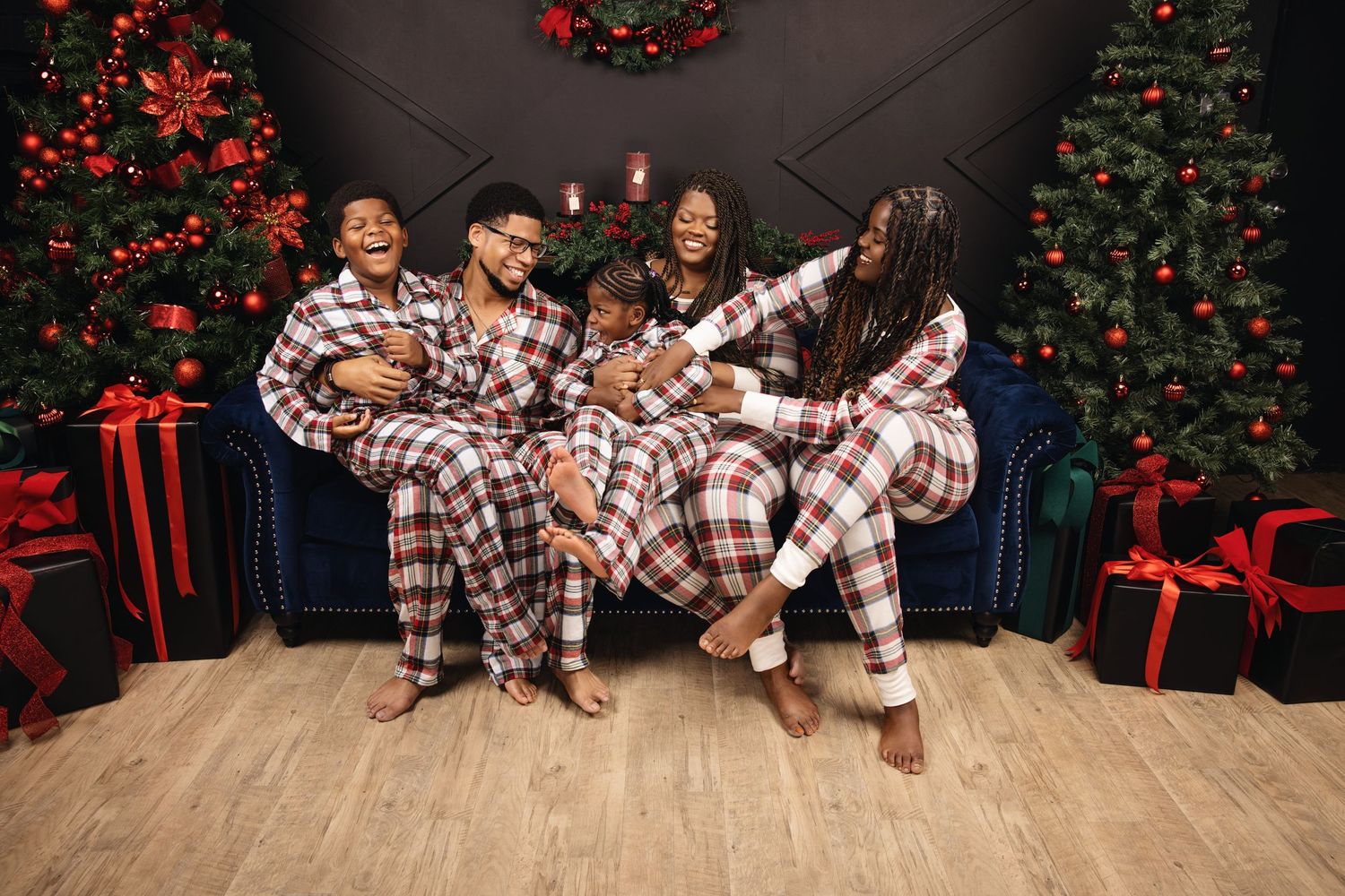 A family in matching plaid pajamas sits together in front of a decorated Christmas tree and holiday backdrop.