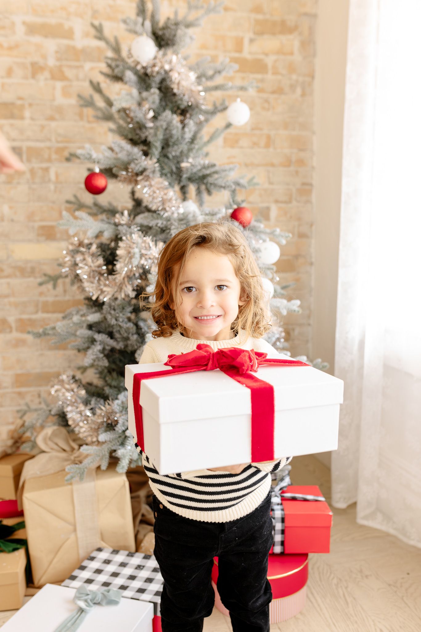 Child holds white wrapped Christmas presents near decorated tree with red ornaments and snowy branches in bright room.