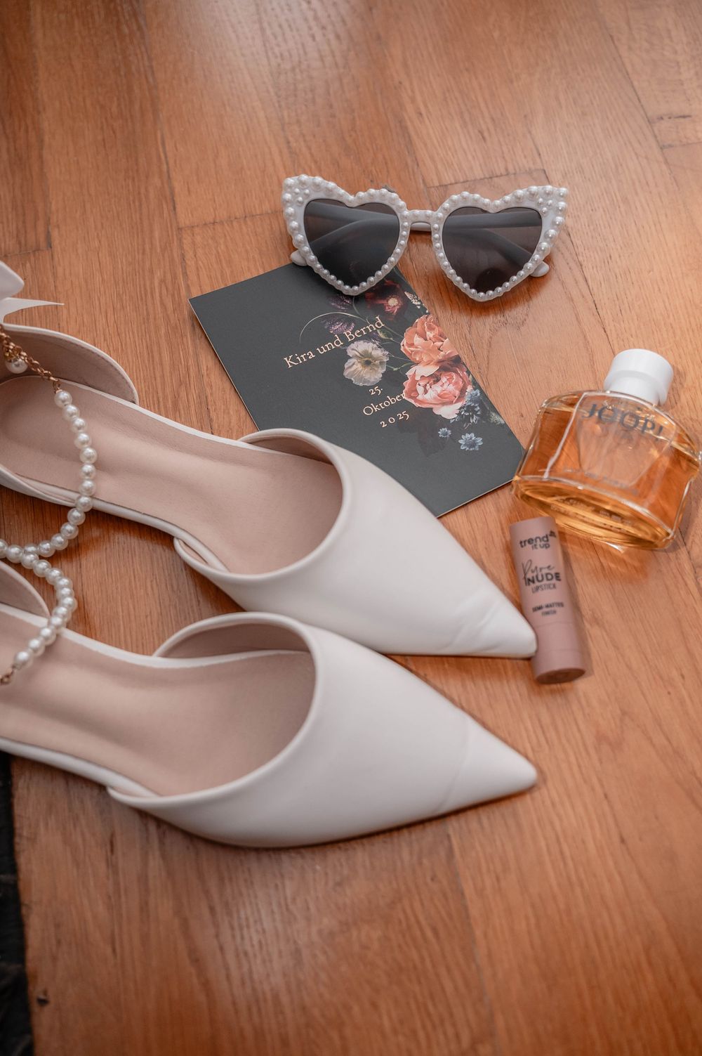 White bridal shoes with pearl accessories and perfume bottle arranged on wooden floor.