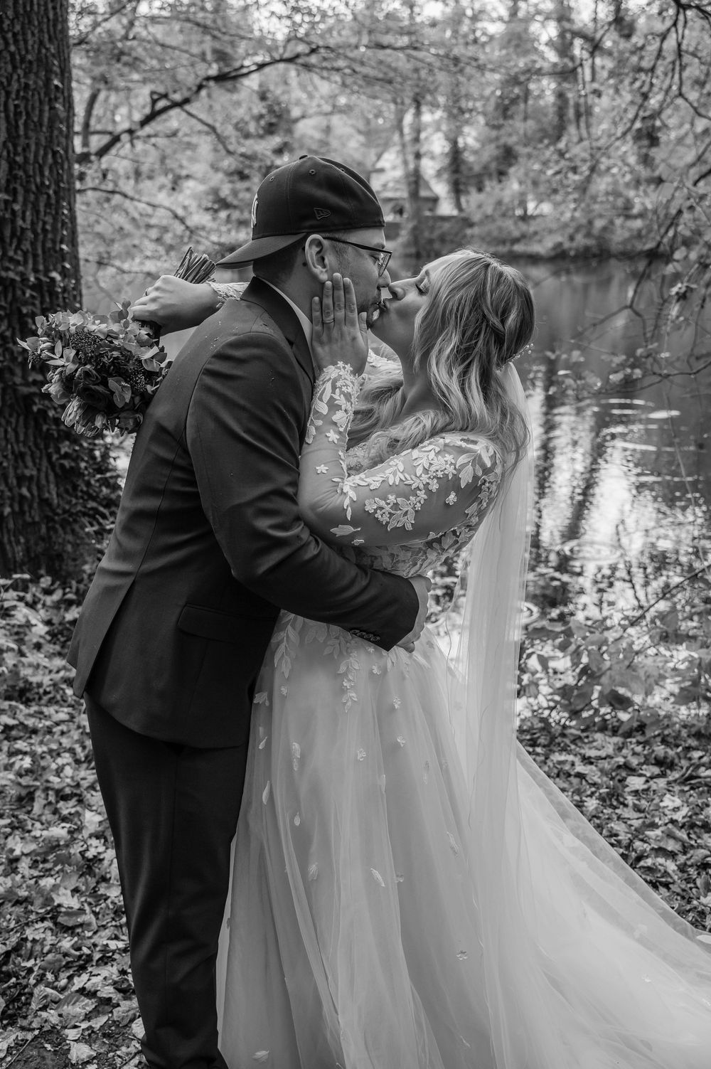 A romantic black and white wedding photo of a couple embracing by a reflective pond in a wooded setting.