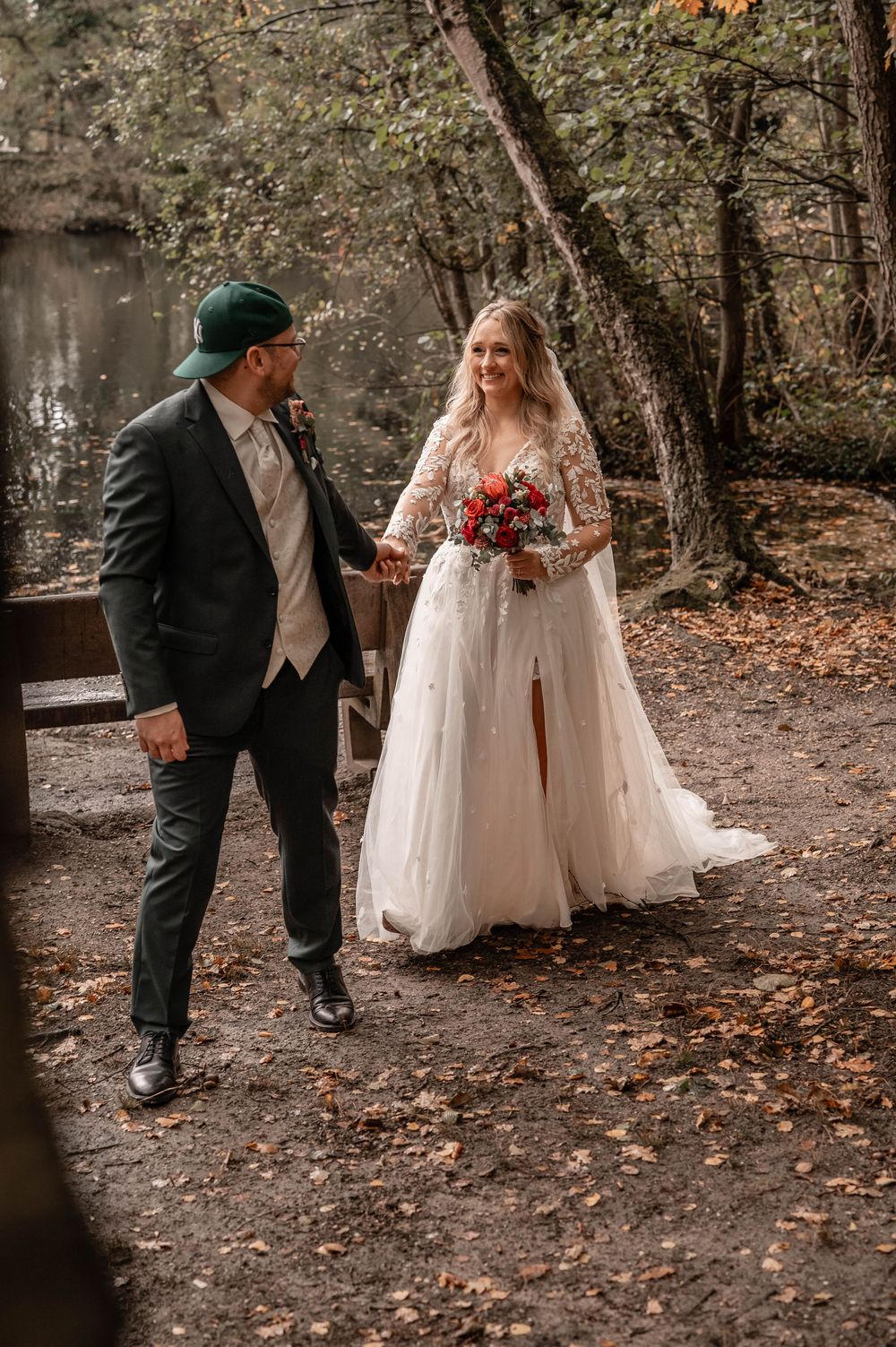 A couple in wedding attire walks through an autumn forest with fallen leaves on the ground.