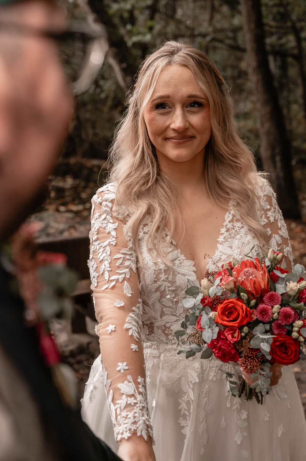 A wedding portrait showing a lace dress and red rose bouquet against a forest backdrop.