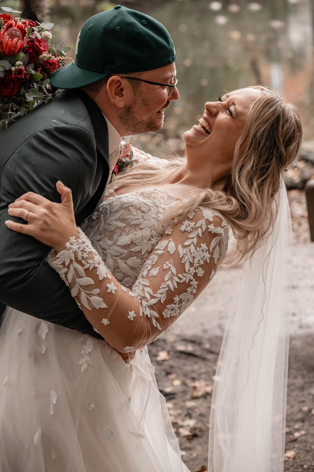 Romantic wedding couple embracing outdoors with bride in lace gown and flowing veil.