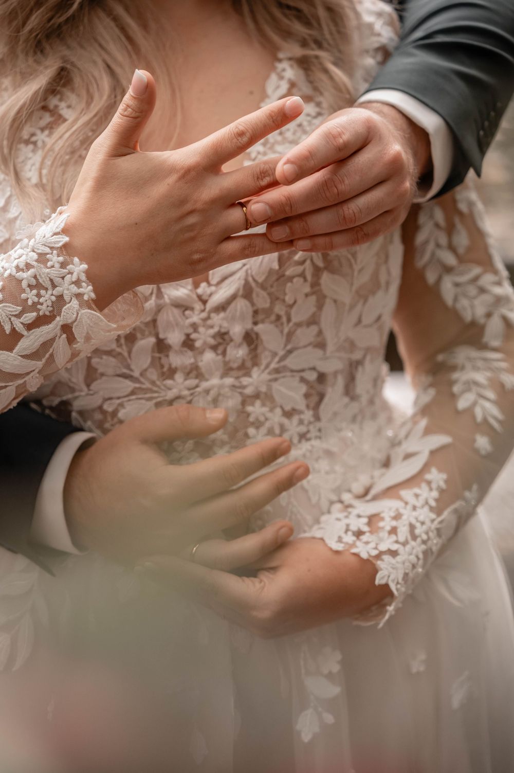 Close-up detail of wedding rings and lace dress sleeves as newlyweds hold hands.