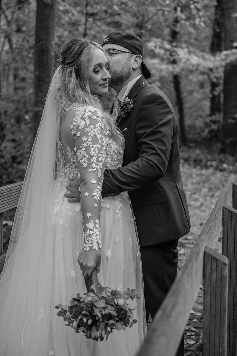 Black and white wedding photo showing tender moment between newlyweds in a forest setting.