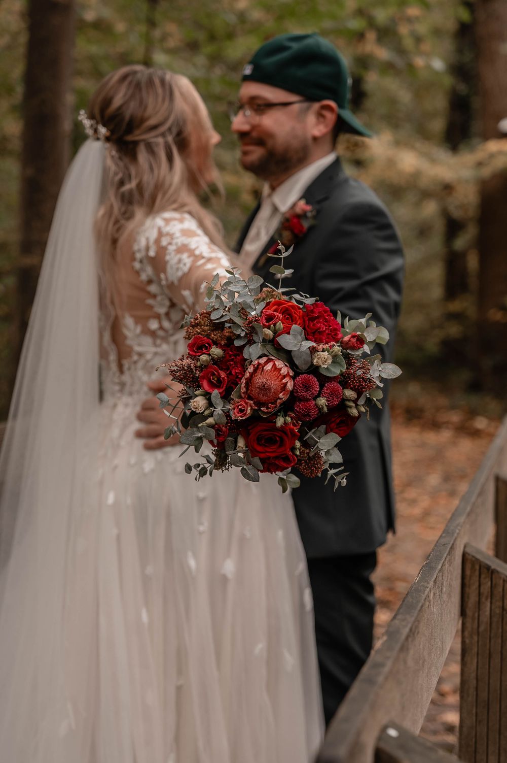 Newlyweds share intimate moment while holding stunning red rose bouquet in autumn forest.