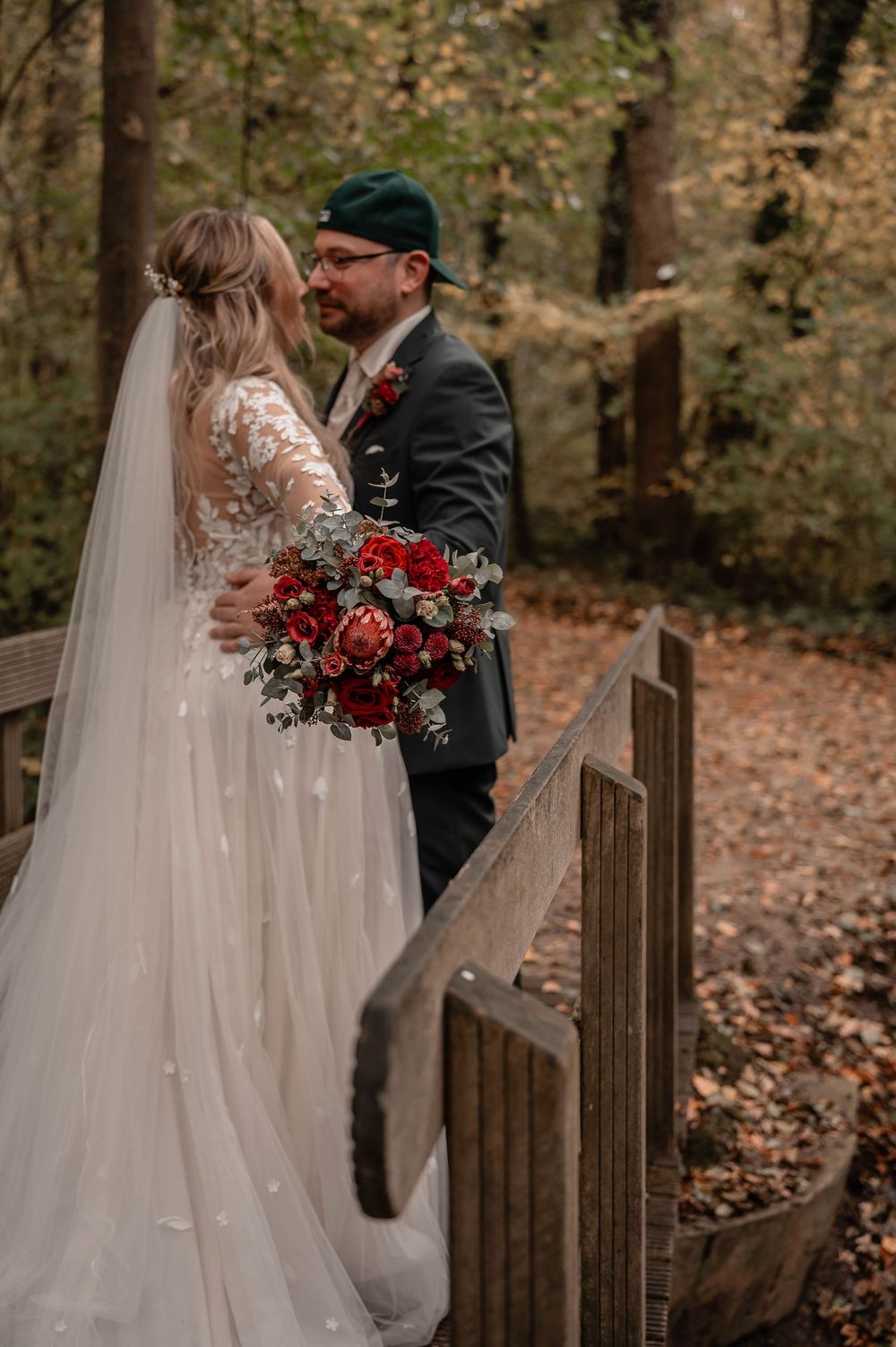 Intimate autumn wedding moment captured on wooden bridge surrounded by fall foliage and red floral bouquet.