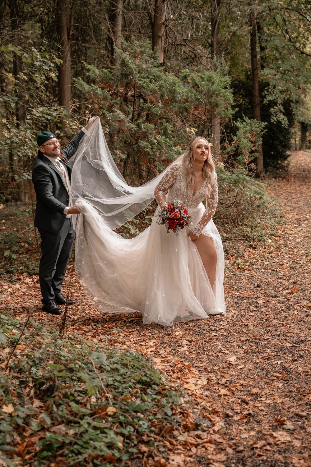 Newlyweds posing in a forest with flowing white wedding dress train spread across autumn leaves.