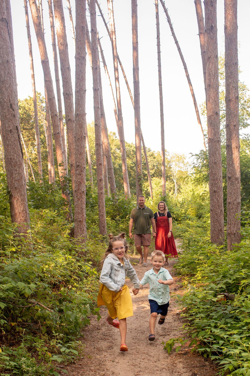Two children running through a forest in front of their parents, smiling happily in Grand Rapids, Michigan