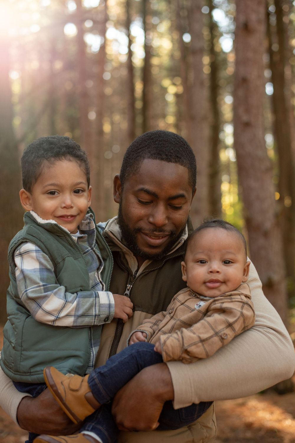 A dad holding his baby and toddler and smiling in front of tall pine trees with the sun shining in Grand Rapids, Michigan