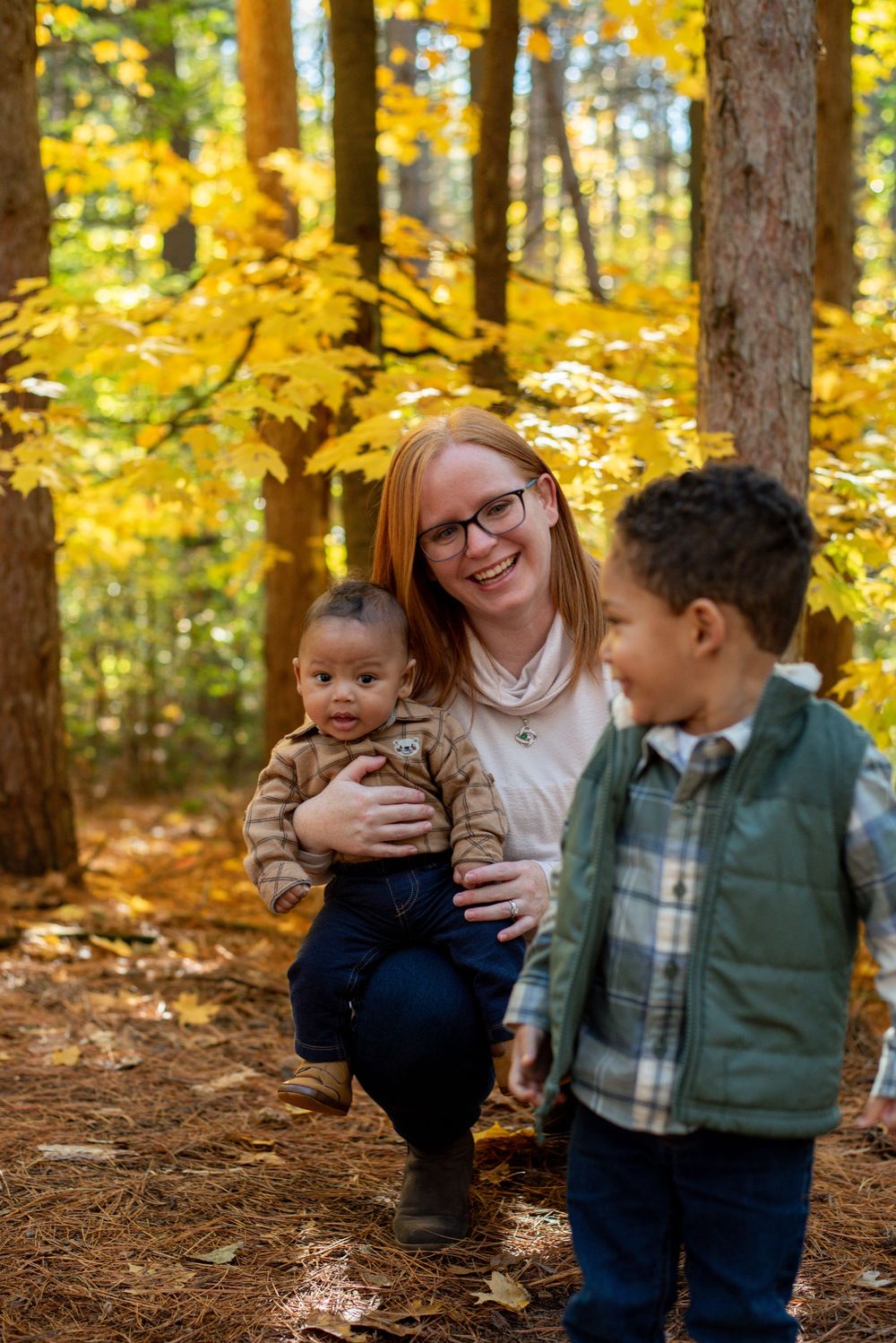 A mom holding her baby and lovingly smiling at her toddler in front of a golden, autumn tree in Grand Rapids, Michigan