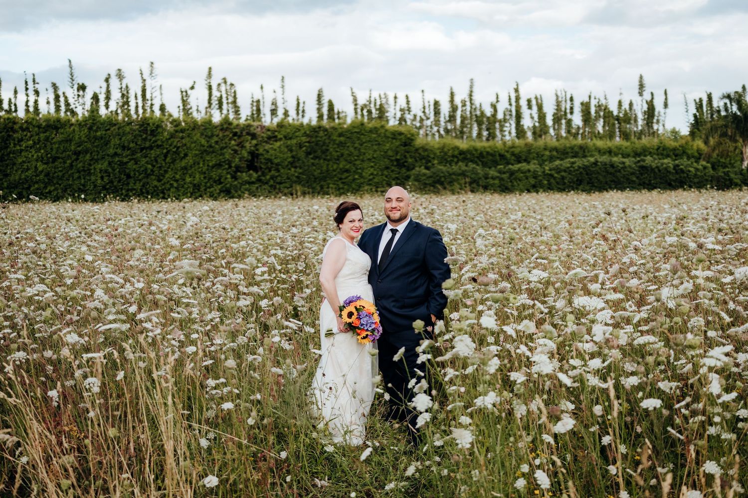 A couple stands together in a dreamy field of white wildflowers with tall green hedges in the background.