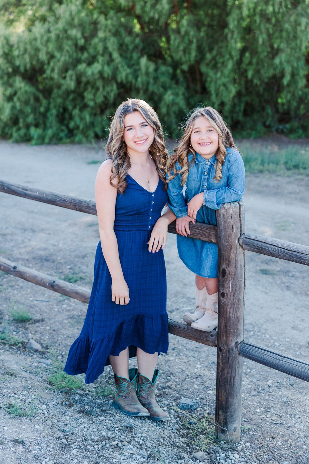 Two friends in denim outfits lean against a rustic wooden fence with green trees in the background.