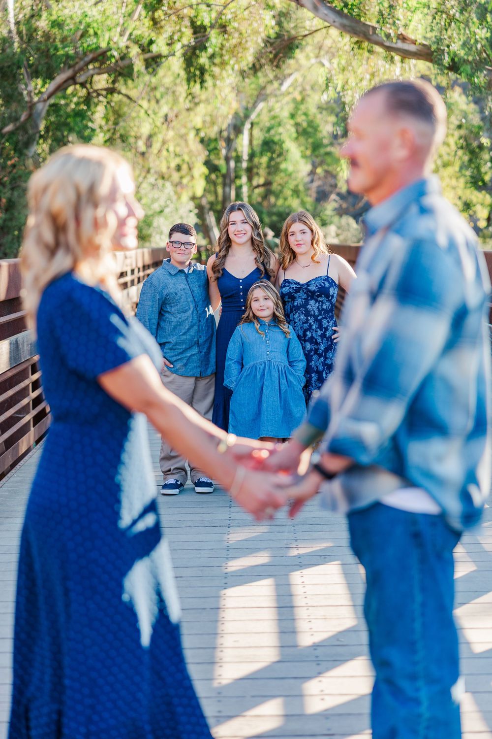 A family wearing coordinating blue outfits poses together on a wooden bridge in a natural outdoor setting.