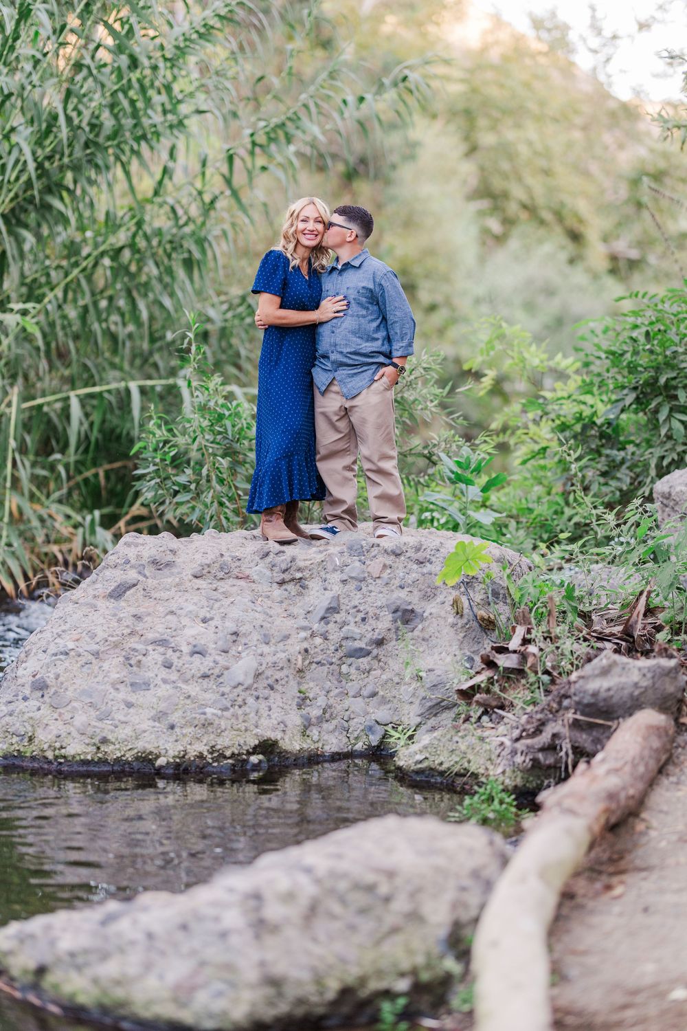 Romantic couple sharing intimate moments by a creek with natural greenery and rocks in the background.