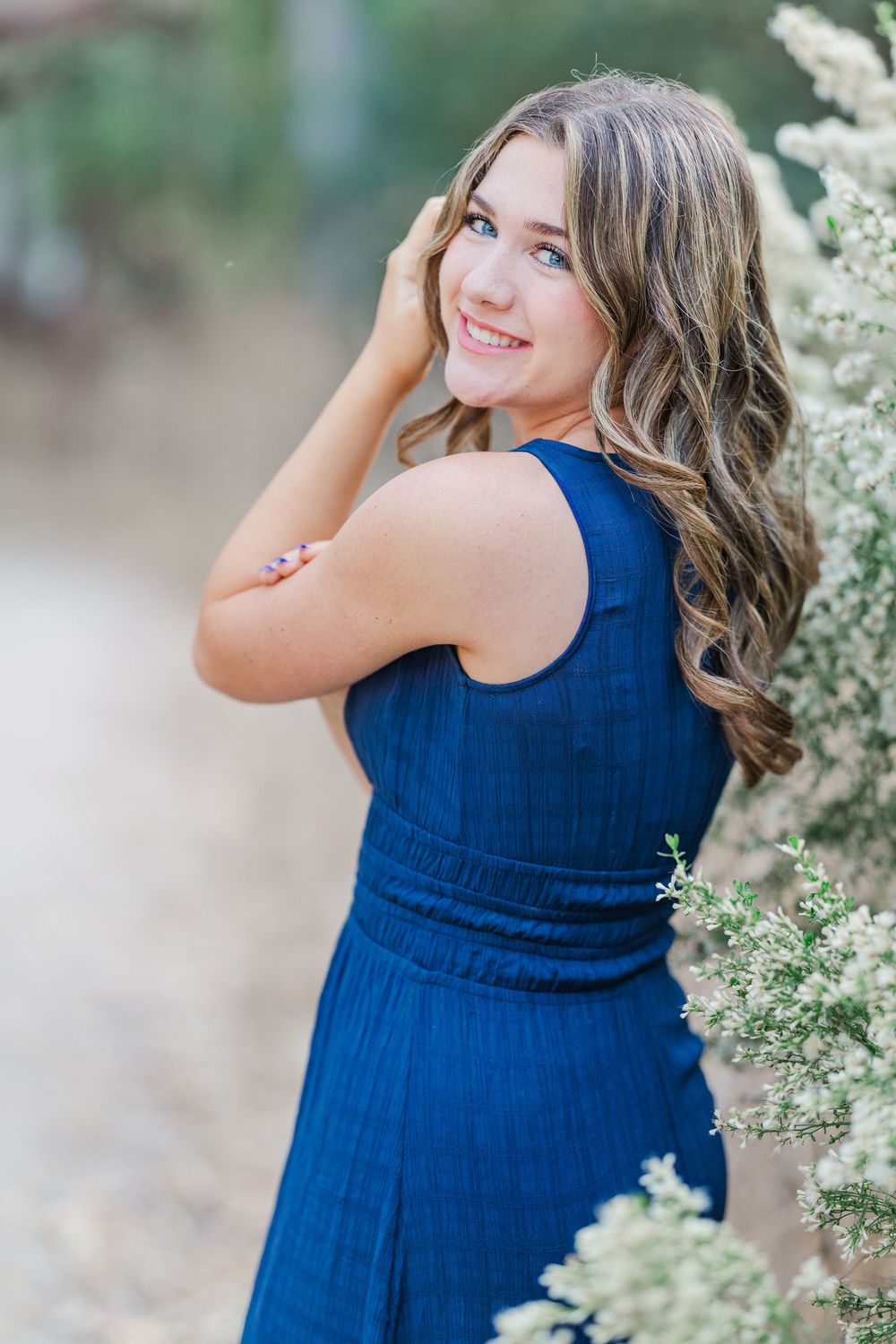 A person in a navy blue dress poses gracefully against a backdrop of delicate white flowers in a garden setting.