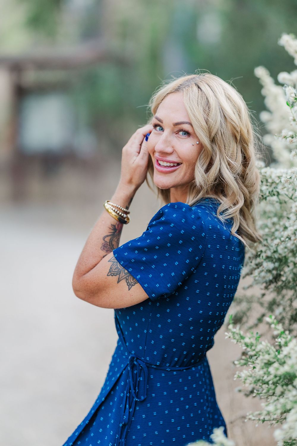 A person in a royal blue polka dot dress poses near blooming white flowers in an outdoor setting.