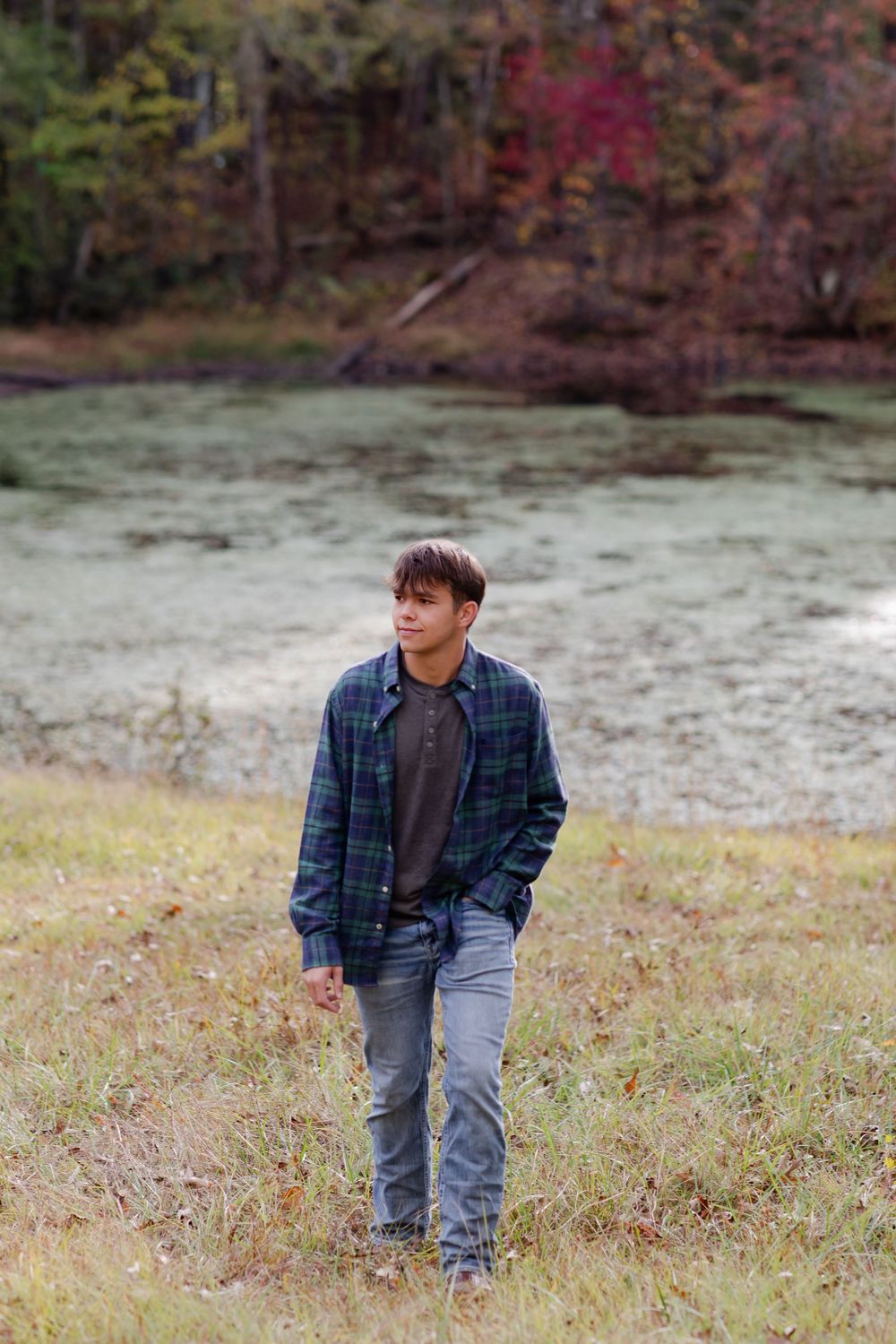 A person wearing a plaid shirt and jeans stands in a grassy autumn field with fall foliage in the background.
