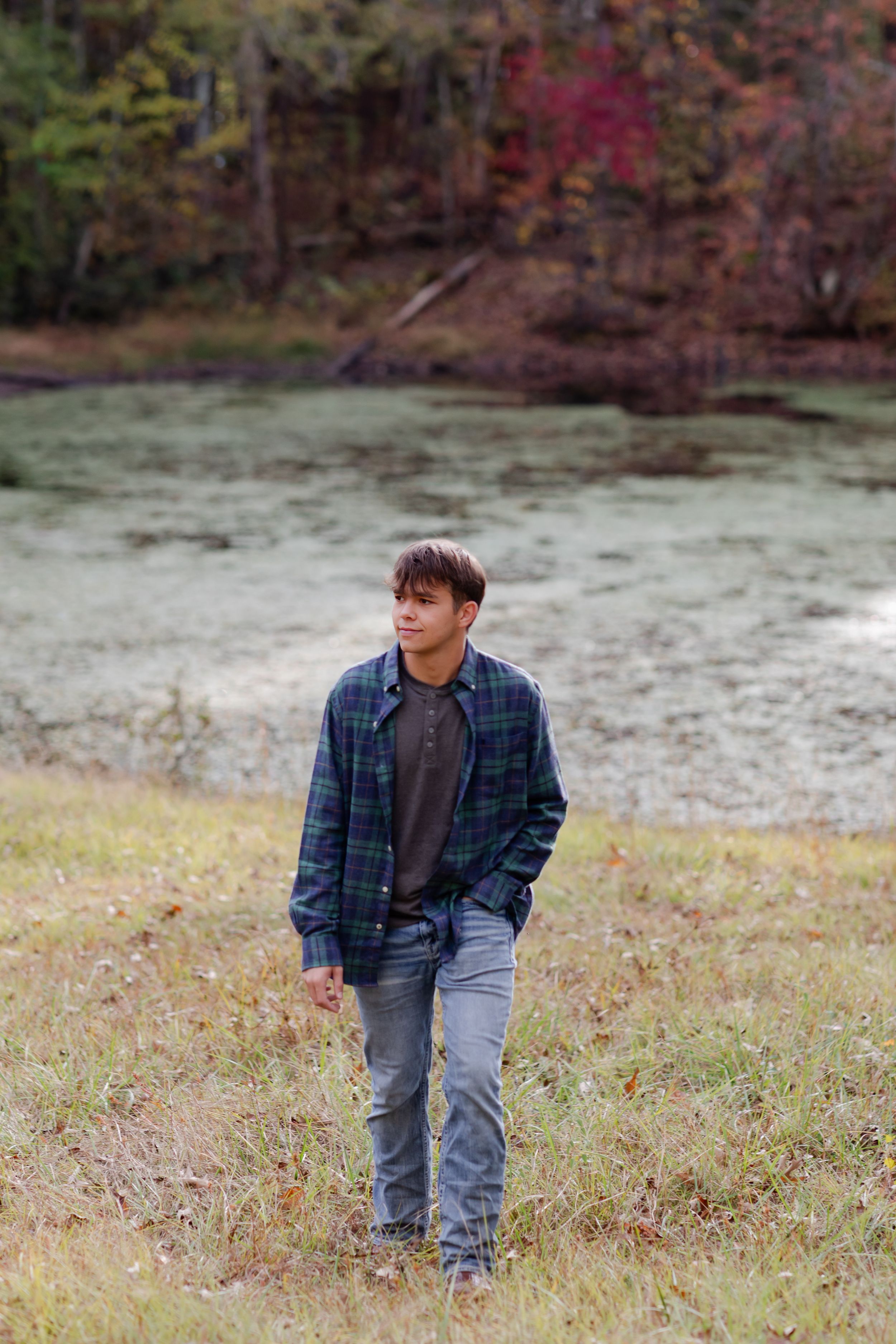A person wearing a plaid shirt and jeans stands in a grassy autumn field with fall foliage in the background.