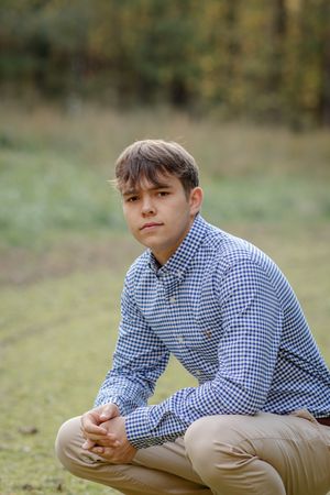 A person wearing a blue and white checkered shirt and khaki pants sits casually on grassy ground outdoors.