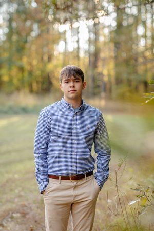 A person in a blue gingham shirt and khaki pants stands in a sunlit forest setting during autumn.