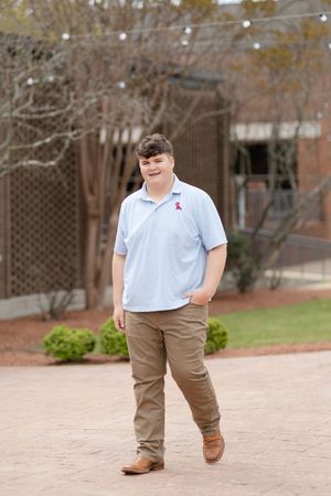 A young person in a light blue polo and khaki pants stands casually outdoors.