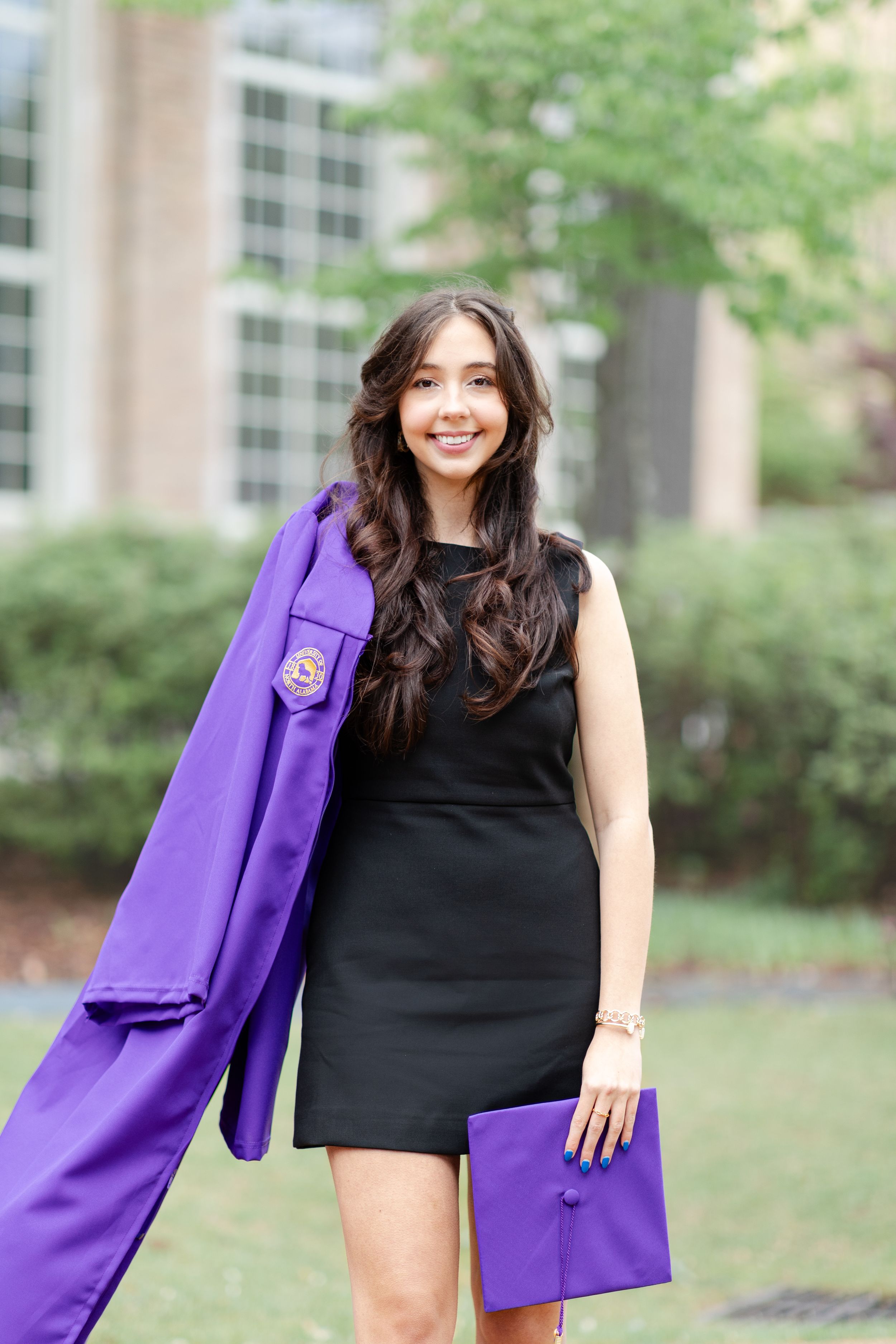 A graduate poses with purple graduation regalia and diploma holder while wearing a black dress.