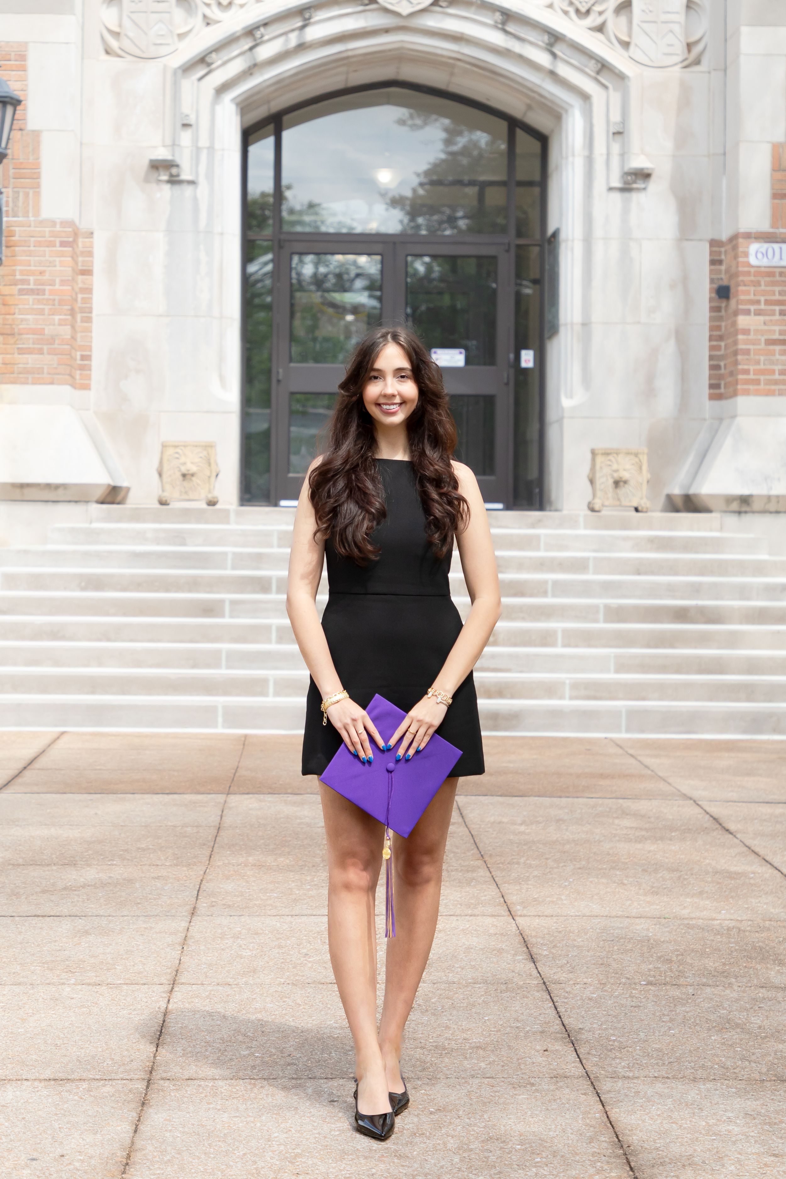 Graduate holding purple diploma folder while posing on steps of brick building.