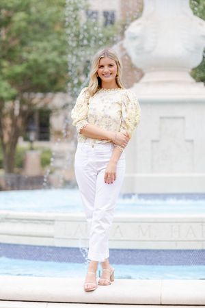 Woman in floral top and white pants poses near fountain on sunny day.
