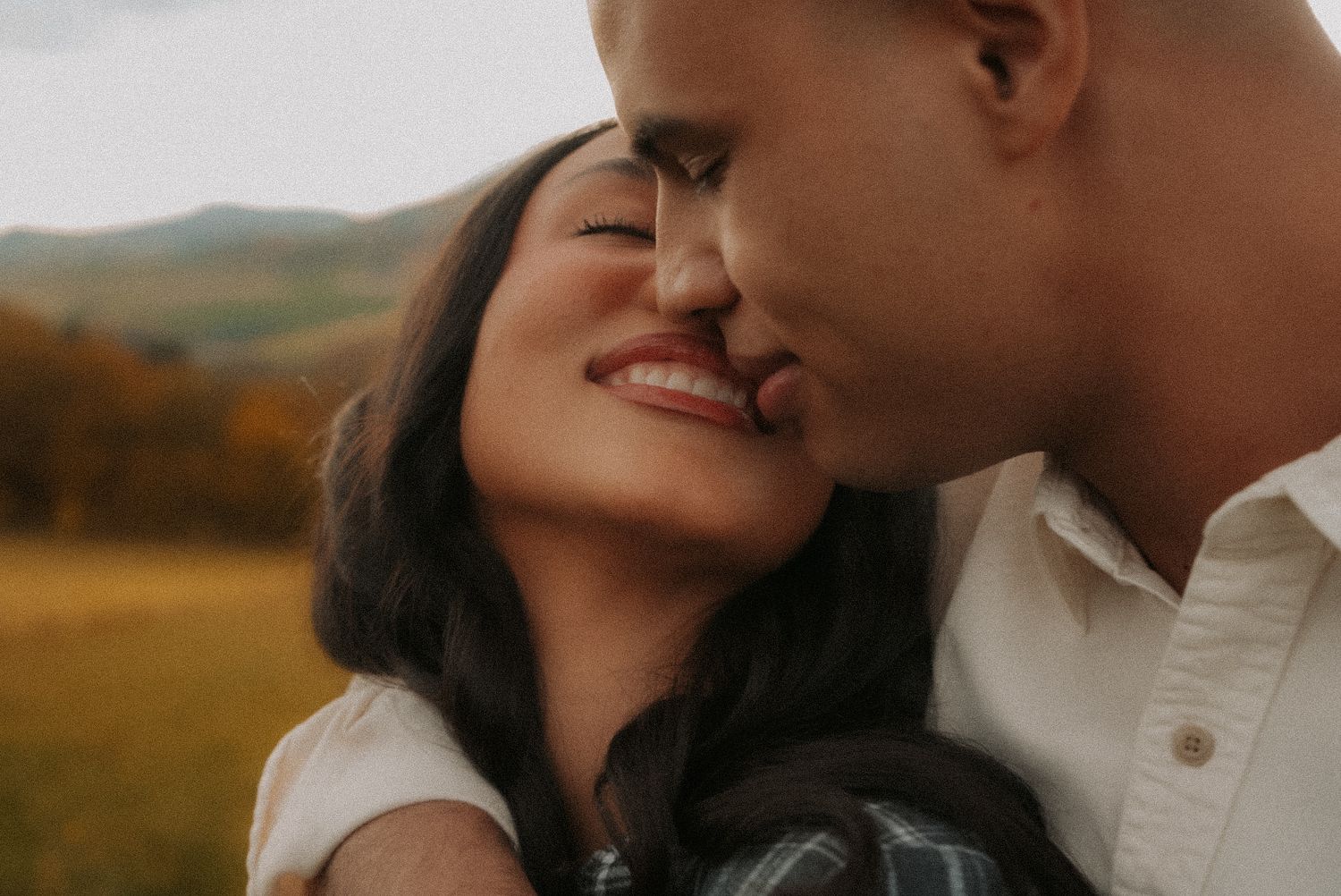A couple shares a romantic moment outdoors with rolling green hills in the background.