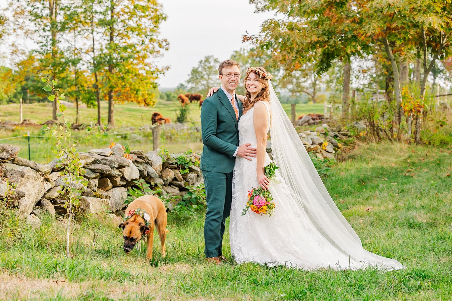 A couple poses with their dog in a rustic outdoor setting during their autumn wedding ceremony.