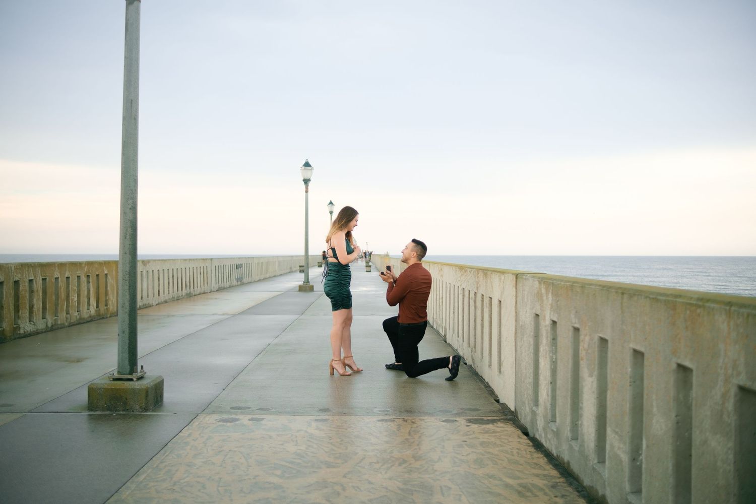 A surprise proposal moment unfolds on an oceanside boardwalk during golden hour.