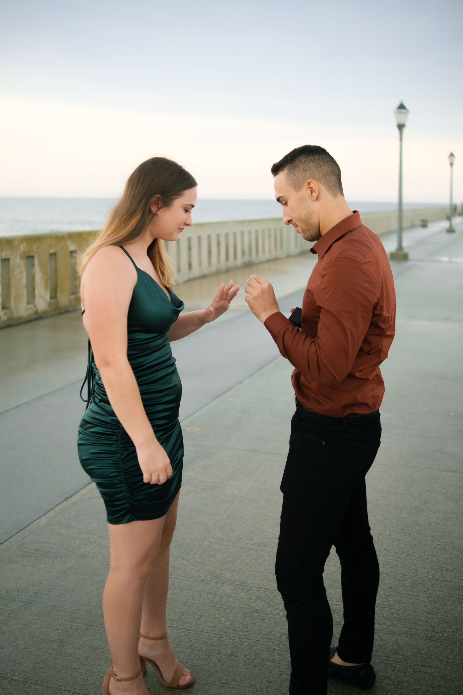A romantic proposal scene on a coastal walkway with a woman in green dress and man in brown sweater.