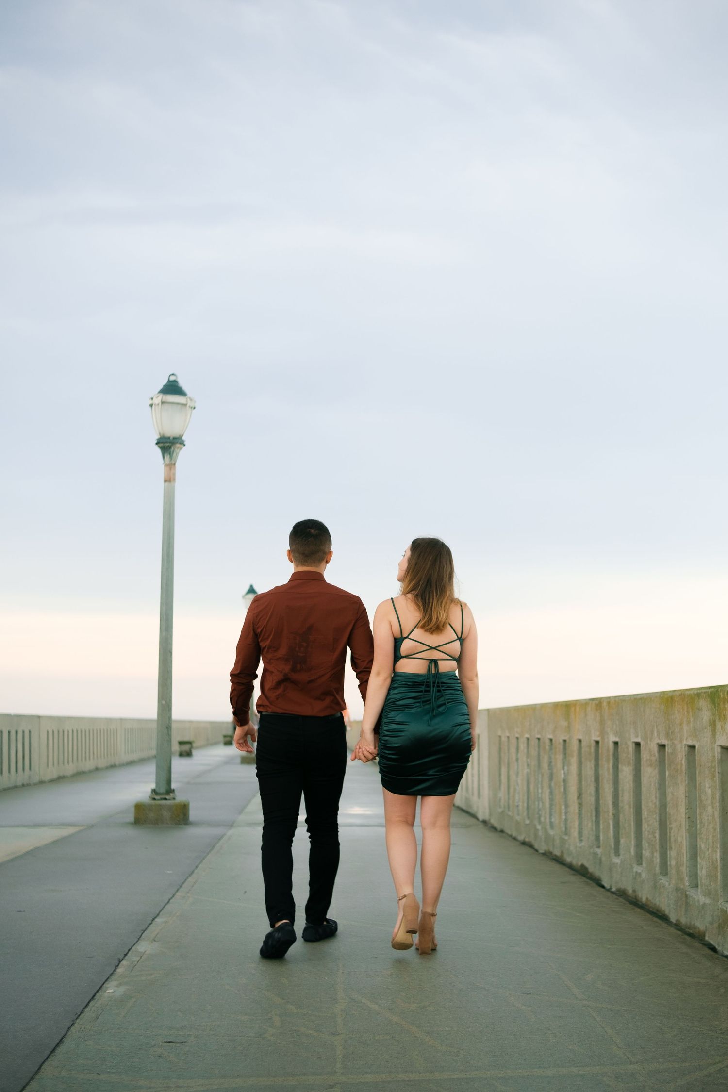 Two people walk hand in hand along a concrete pier with lampposts and ocean views.