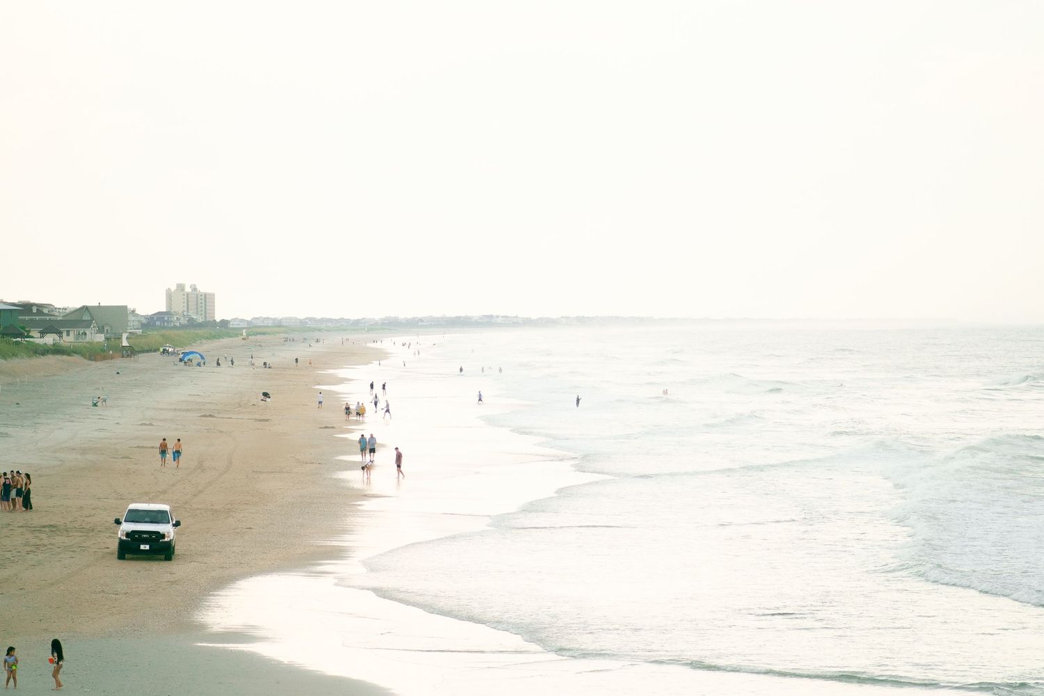 A wide view of a beach shoreline with scattered people and a vehicle visible in the distance.