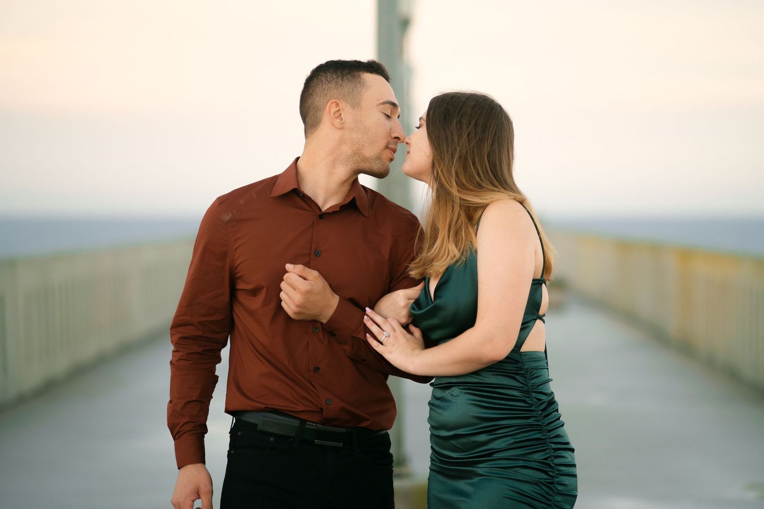 A romantic couple in formal attire sharing an intimate moment on a pier at sunset.