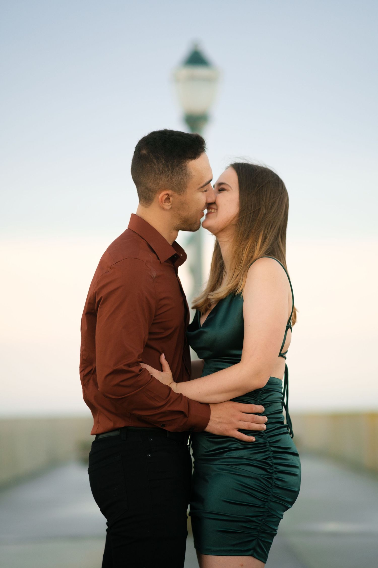 A romantic couple shares an intimate moment on a pier at sunset wearing burgundy and emerald outfits.