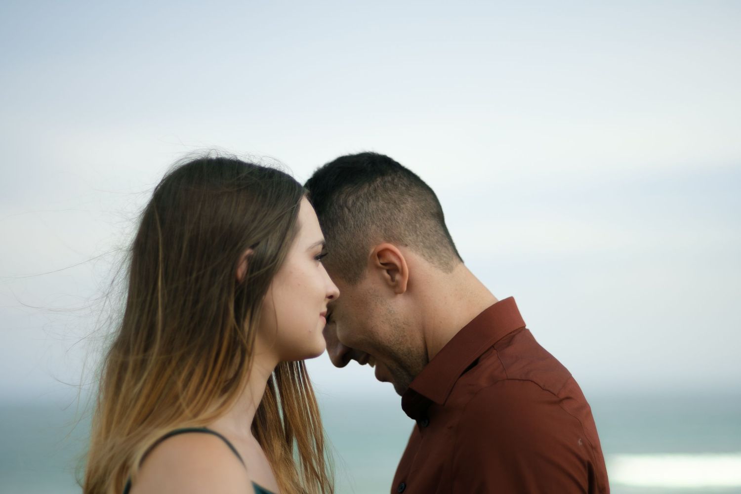 A romantic couple shares an intimate moment against a cloudy sky backdrop on a seaside walkway.