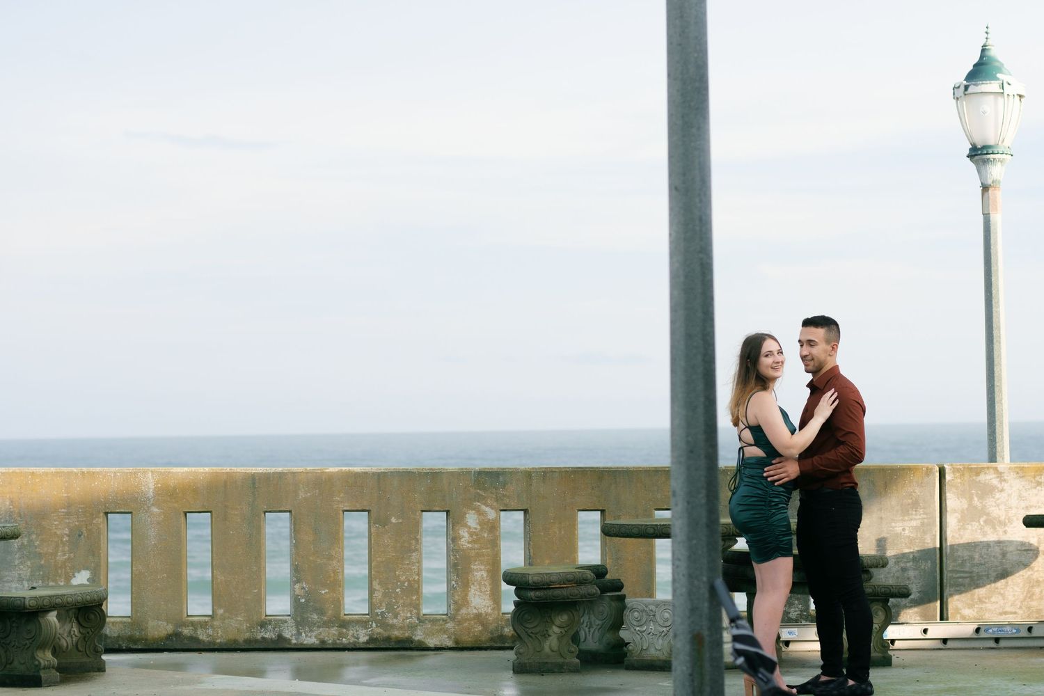 A couple shares an intimate moment on a seaside boardwalk with an ornate lamp post and railing at sunset.