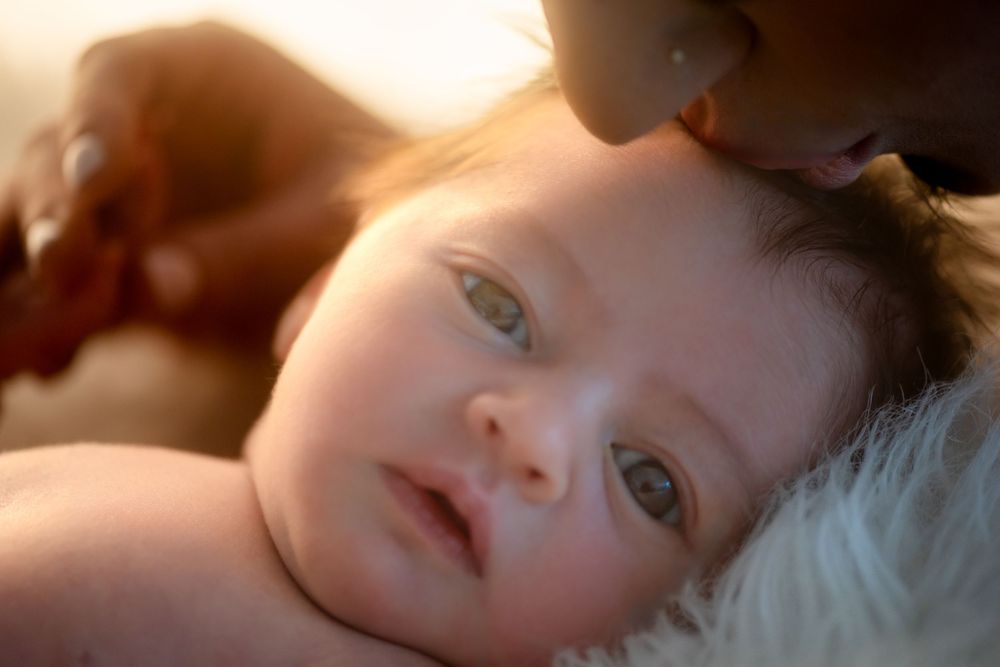 A newborn baby with blue eyes lying on a white fluffy blanket while a parent gently kisses their head.