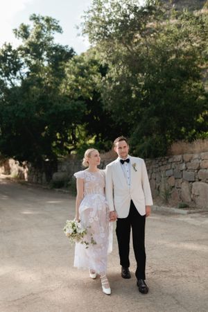 A couple strolls down a sunlit path in formal wedding attire surrounded by stone walls and greenery.