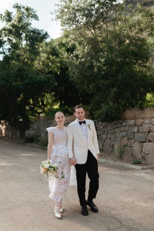 A couple in wedding attire walk hand in hand along a stone-walled path in an outdoor garden setting.